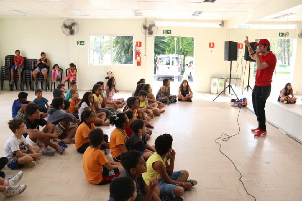 Aula de música para crianças, onde o professor está de pé e as crianças sentadas diante dele
